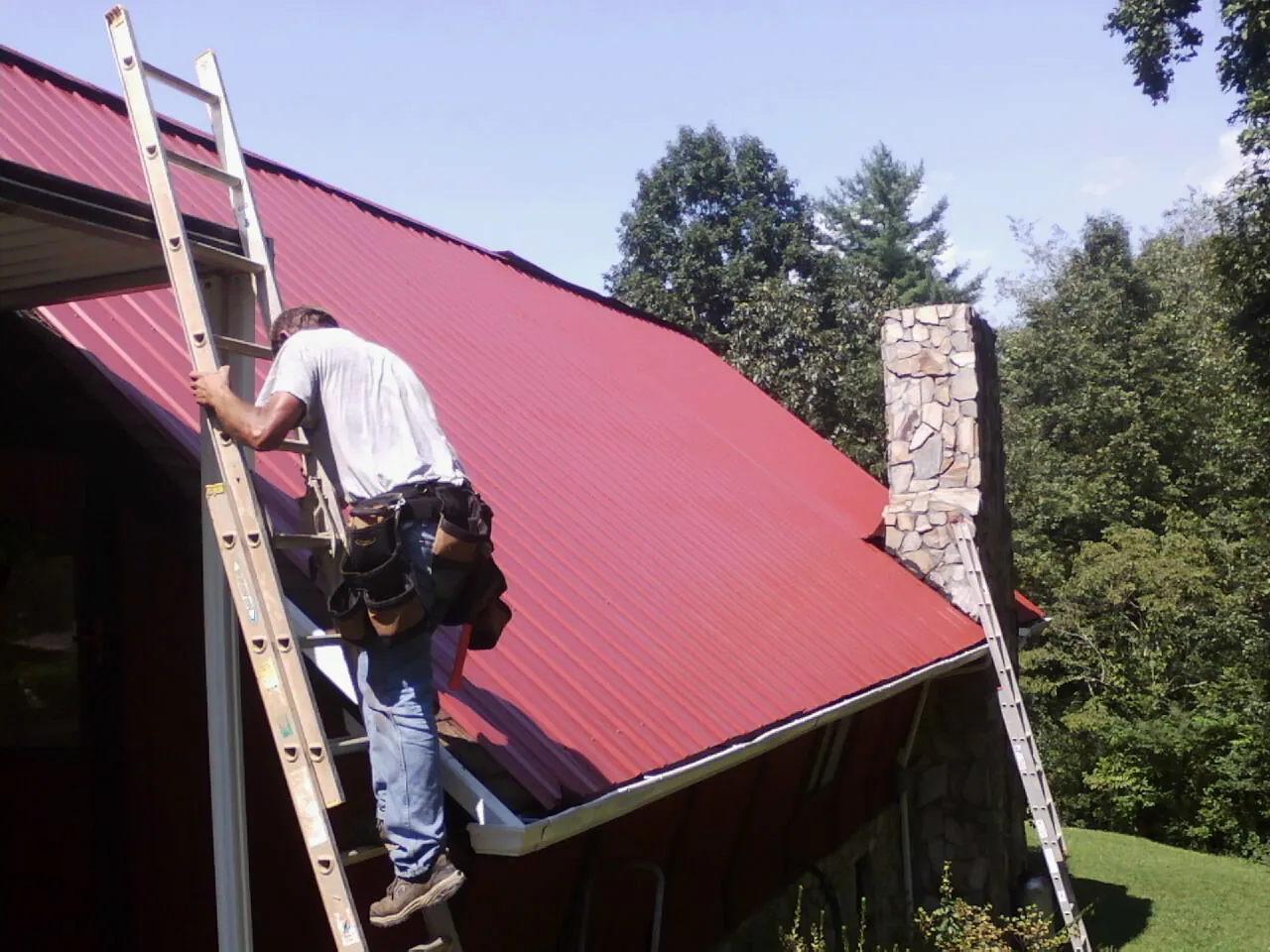 Roofer on a ladder beside a red metal roof and stone chimney — Soffit & Fascia Repair by Roofing Brothers in Hines